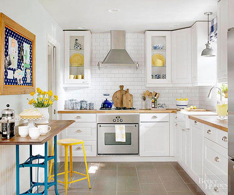 This white kitchen has grey tile floors and wood countertops.  It is accented with a yellow stool and plates, along with blue table legs and dishes.