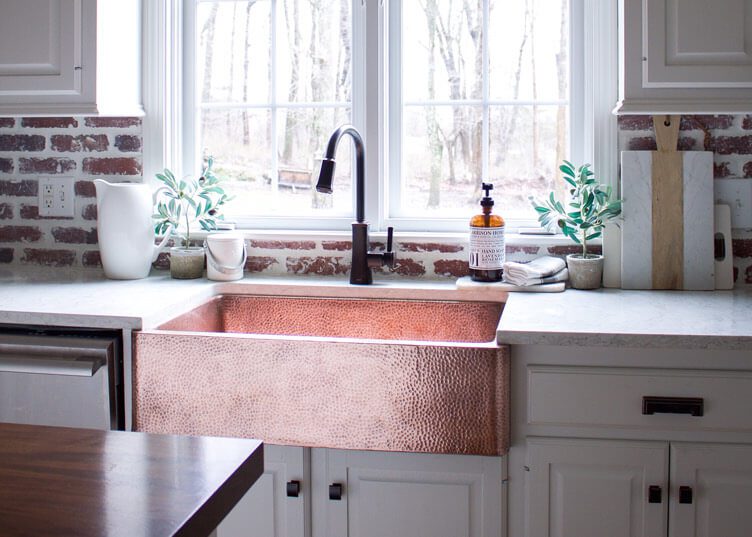 A copper sink that stands out against white cabinets and a brick wall.