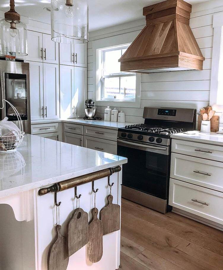 Four wooden cutting boards hang from a rolling pan rack. A wooden vent hood also helps offset all the white.