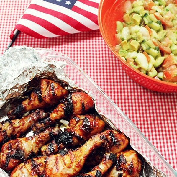 A dish of Fourth of July barbeque chicken wrapped in foil placed next to an American flag and an avocado cucumber salad