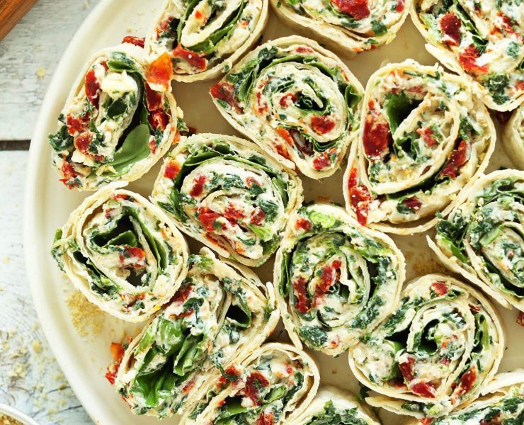A collection of sun-dried tomato and basil pinwheels placed decoratively on a plate, surrounded by loose basil leaves and vegan parmesan cheese crumbles