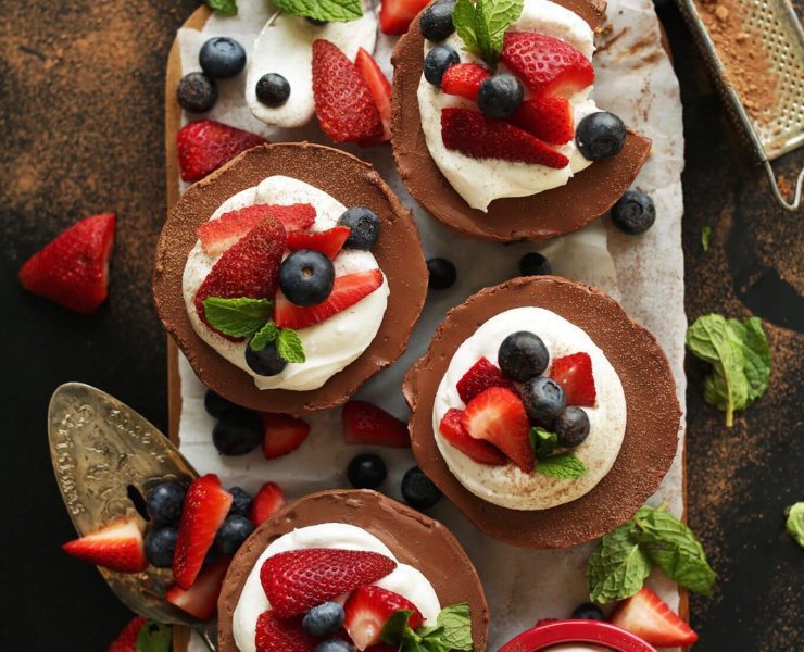 A wooden board complete with mini chocolate cheesecakes topped with coconut whipped cream, strawberries, blueberries, and mint