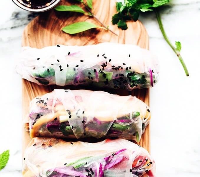 Row of four rainbow summer rolls on a wooden board surrounded by decorative thai basil leaves