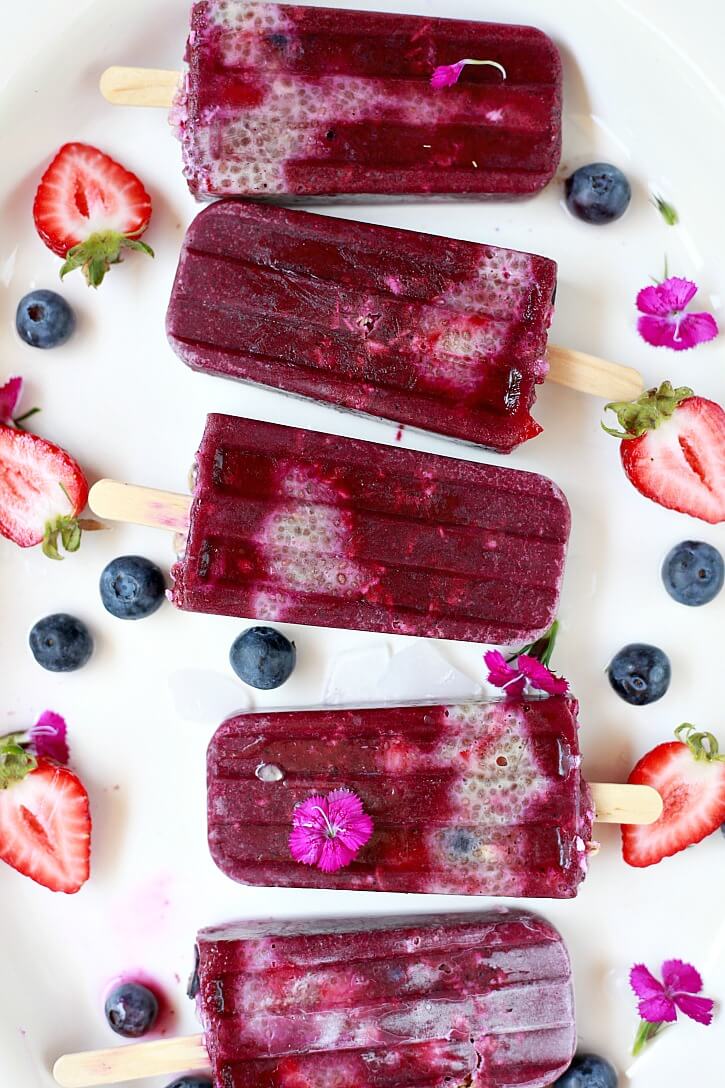 An acai breakfast popsicle family surrounded by an array of diced strawberries, blueberries, and purple flowers