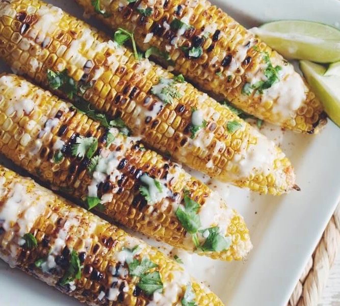 A row of Fourth of July ears of corn drizzled with a coconut lime cream sauce and cilantro, all surrounded by loose lime wedges