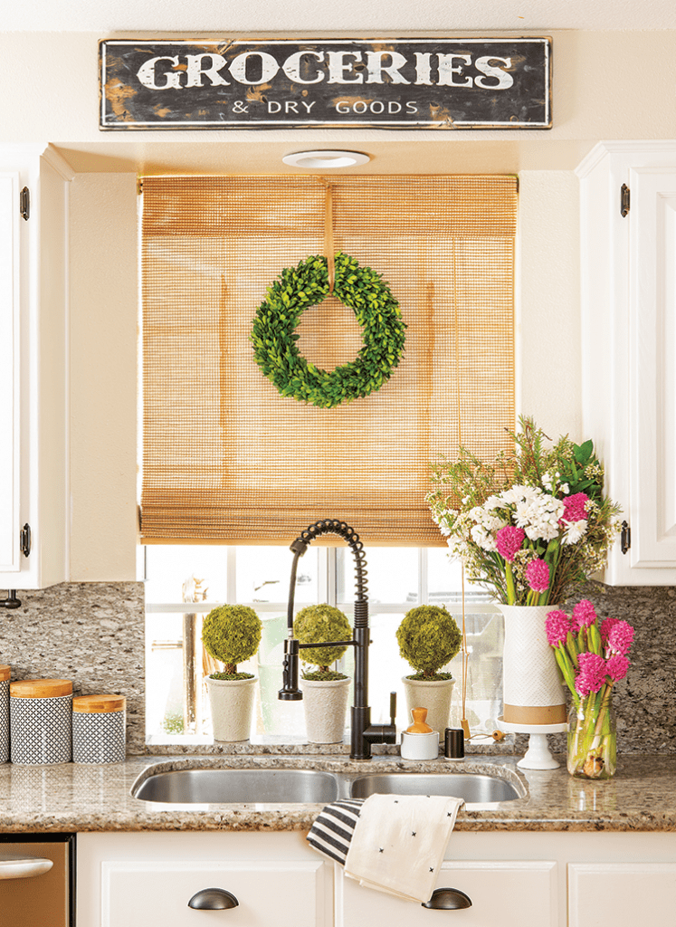 Kitchen sink below a window surrounded by a custom sign, greenery, and farmhouse details.