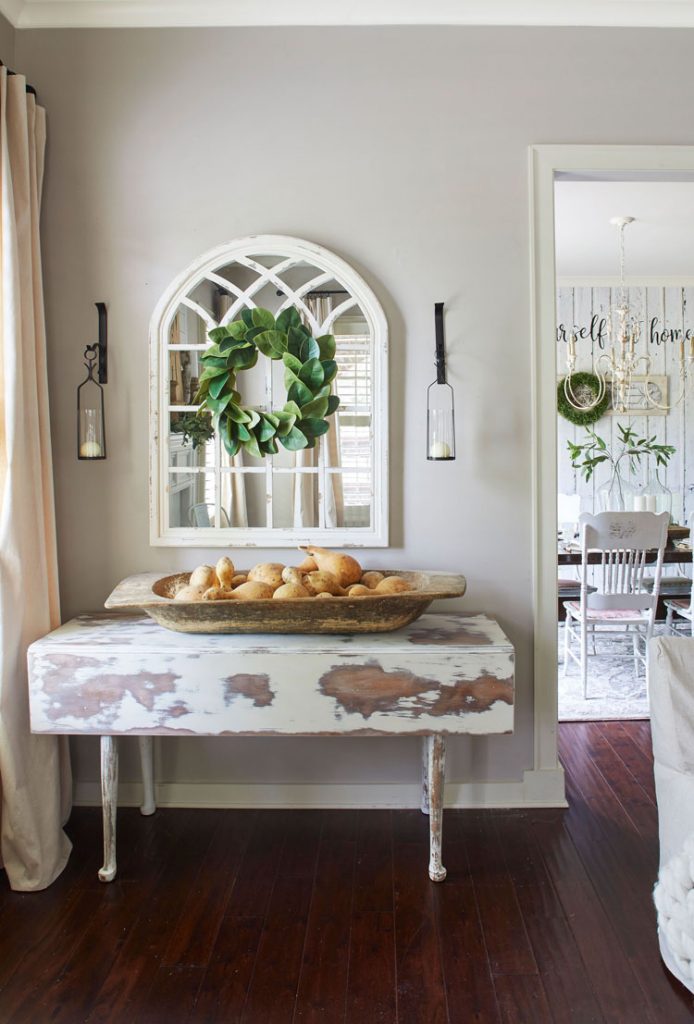 Gourds piled in a rustic wooden bowl on top of a faded white vintage side table.