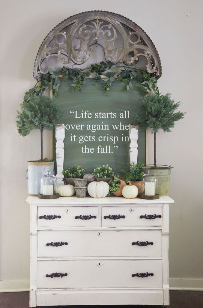 Green chalk board with potted fall plants and white pumpkins on top of an ecru dresser.