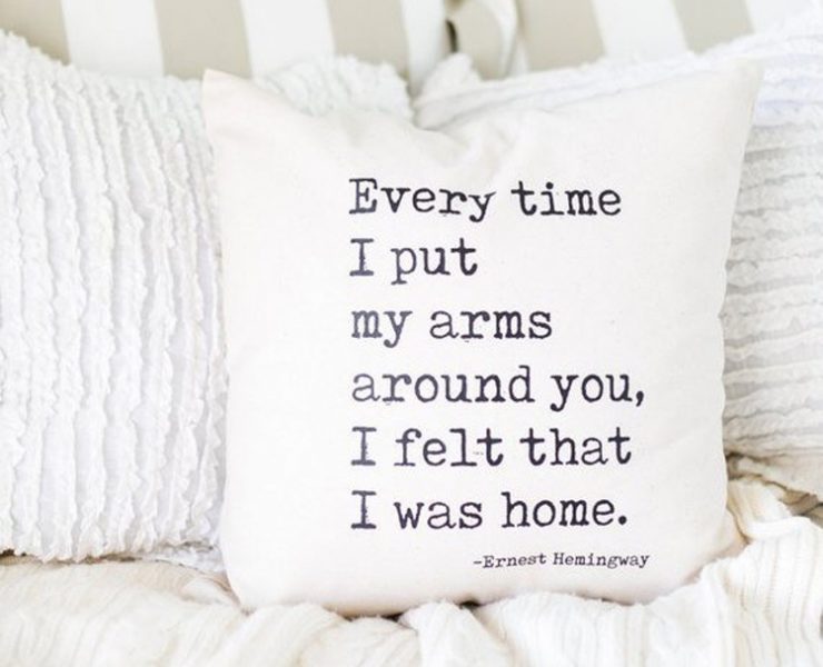 Off-white pillow with black typewriter font that reads "Every time I put my arms around you, I felt that I was home" - Ernest Hemingway.