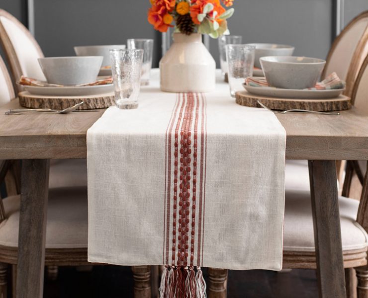 Ivory cotton table runner with rust-colored stripes down the middle and knotted fringe detailing at the ends.
