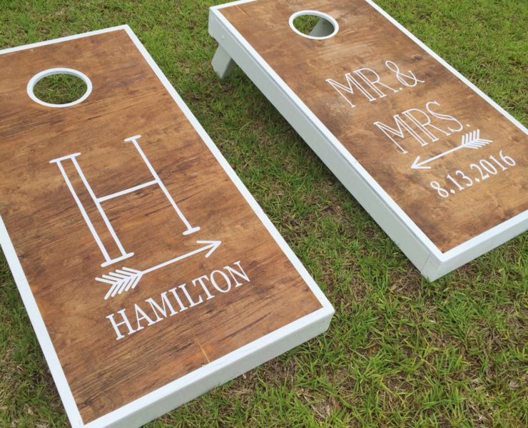 Two cornhole game boards next to each other with white decals and arrows that say “H Hamilton” on the left one and “Mr. and Mrs. (8.13.2016)” on the right one.