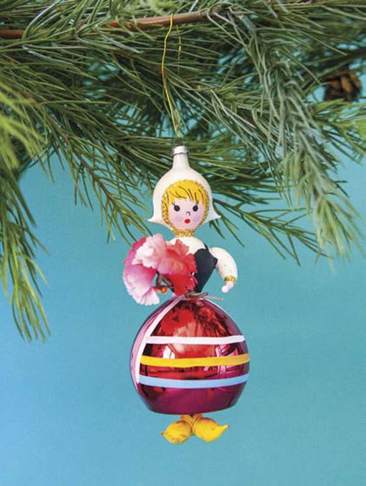 This picture shows the beauty of high-quality collectible Christmas ornaments. A brightly-colored glass ornament in the form and fashion of a peasant Dutch girl hangs from a fir Christmas tree branch.