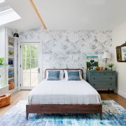 This farm-cottage bedroom shows off the comforts of home with a white comforter, peel-away gray patterned wallpaper and plenty of windows for natural light.