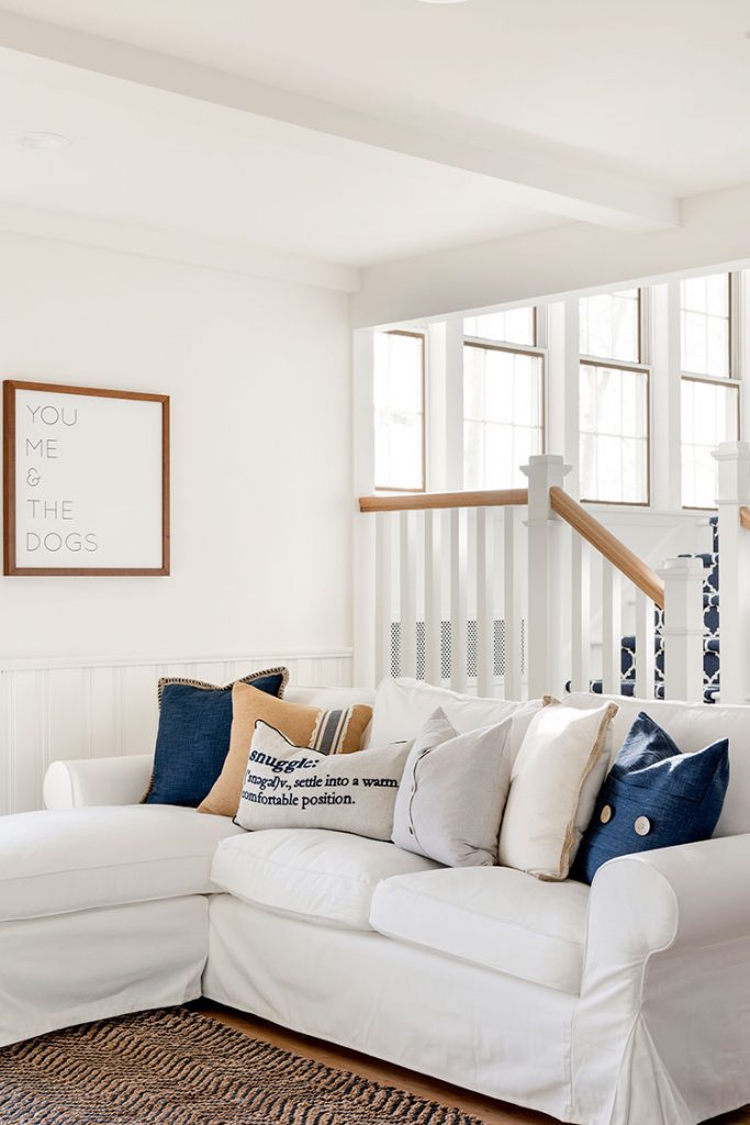 The white living room couch next to the stairwell entrance has six unique pillows in a neutral and blue color palette. The wall art above it says "You, Me & The Dogs".