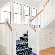 The farmhouse stairway has a dark blue and white patterned carpet with four large paneled windows in the wall next to it.