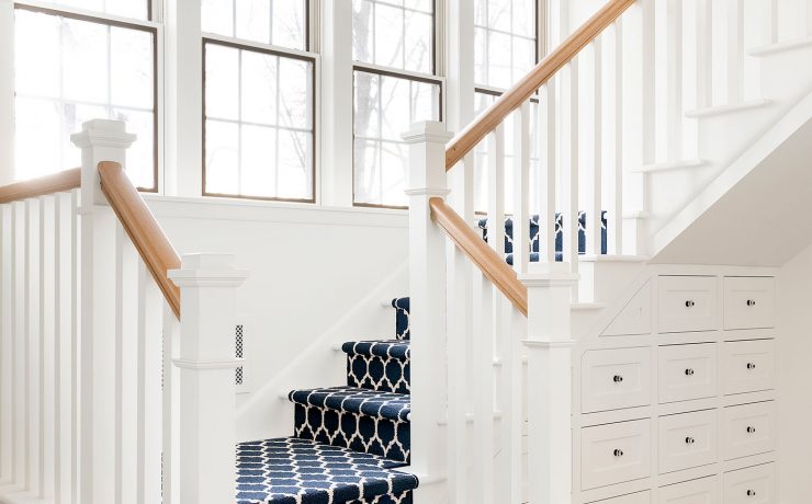 The farmhouse stairway has a dark blue and white patterned carpet with four large paneled windows in the wall next to it.