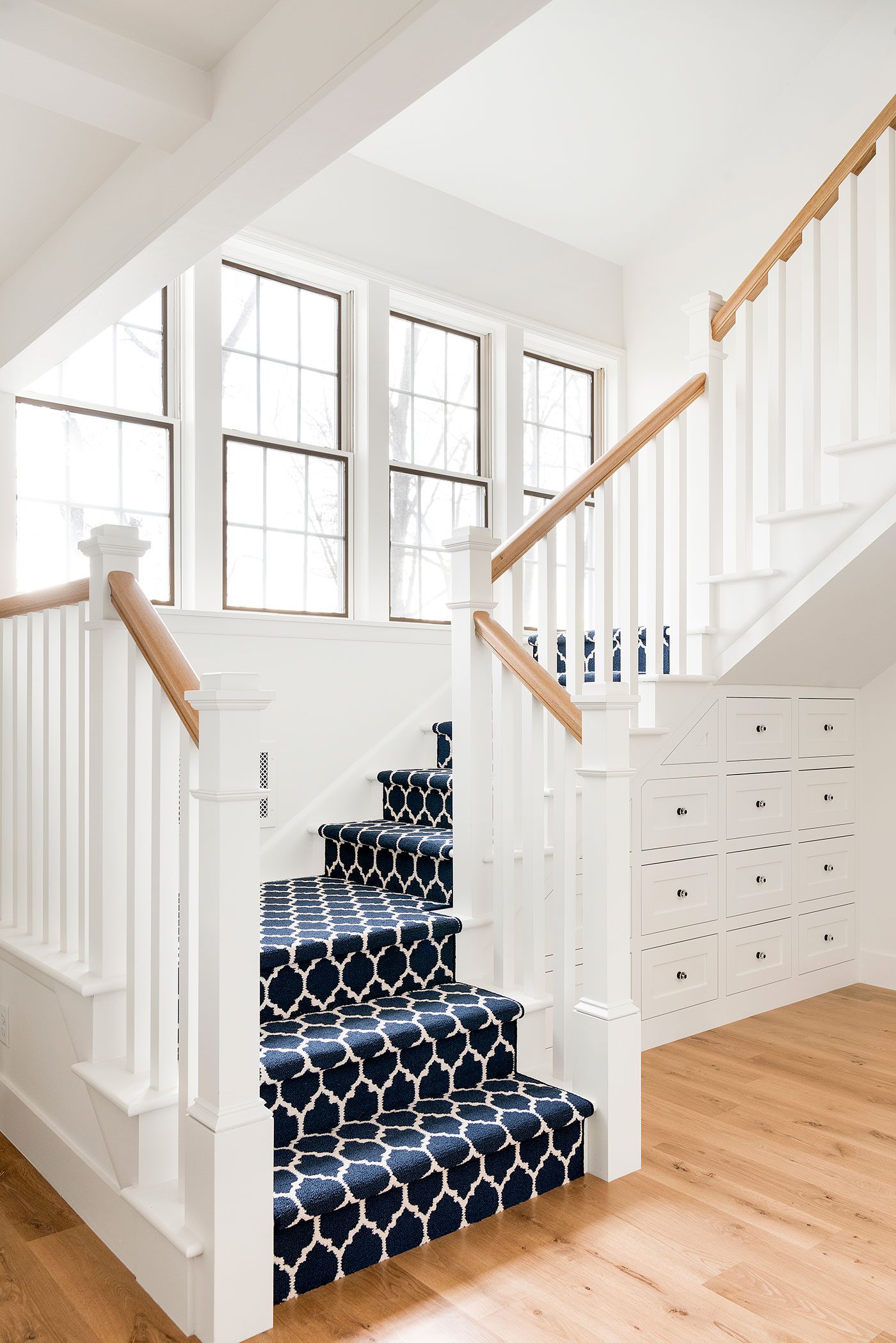 The farmhouse stairway has a dark blue and white patterned carpet with four large paneled windows in the wall next to it.