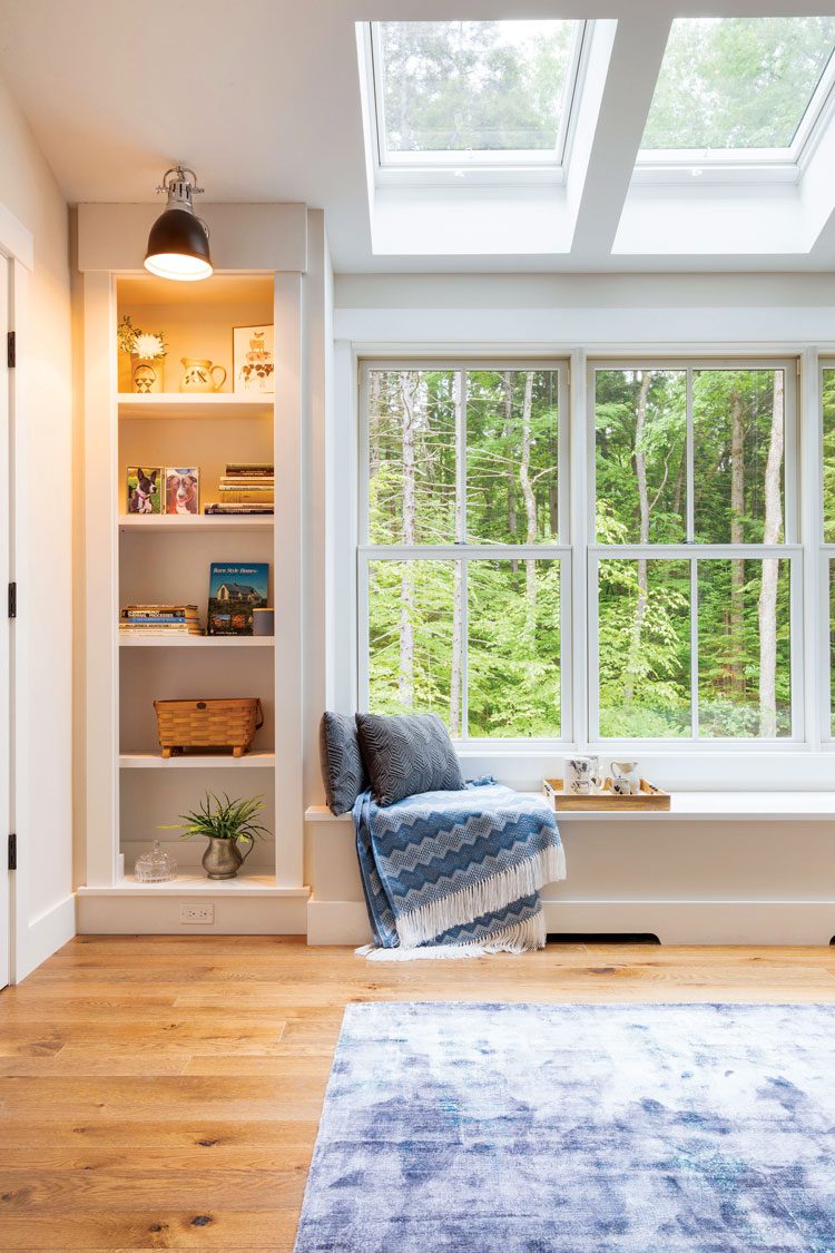 This farm-cottage bedroom window seat is below ceiling windows as well as beside wall windows. It has two gray pillows and a blue blanket and built in wall-shelves next to it.