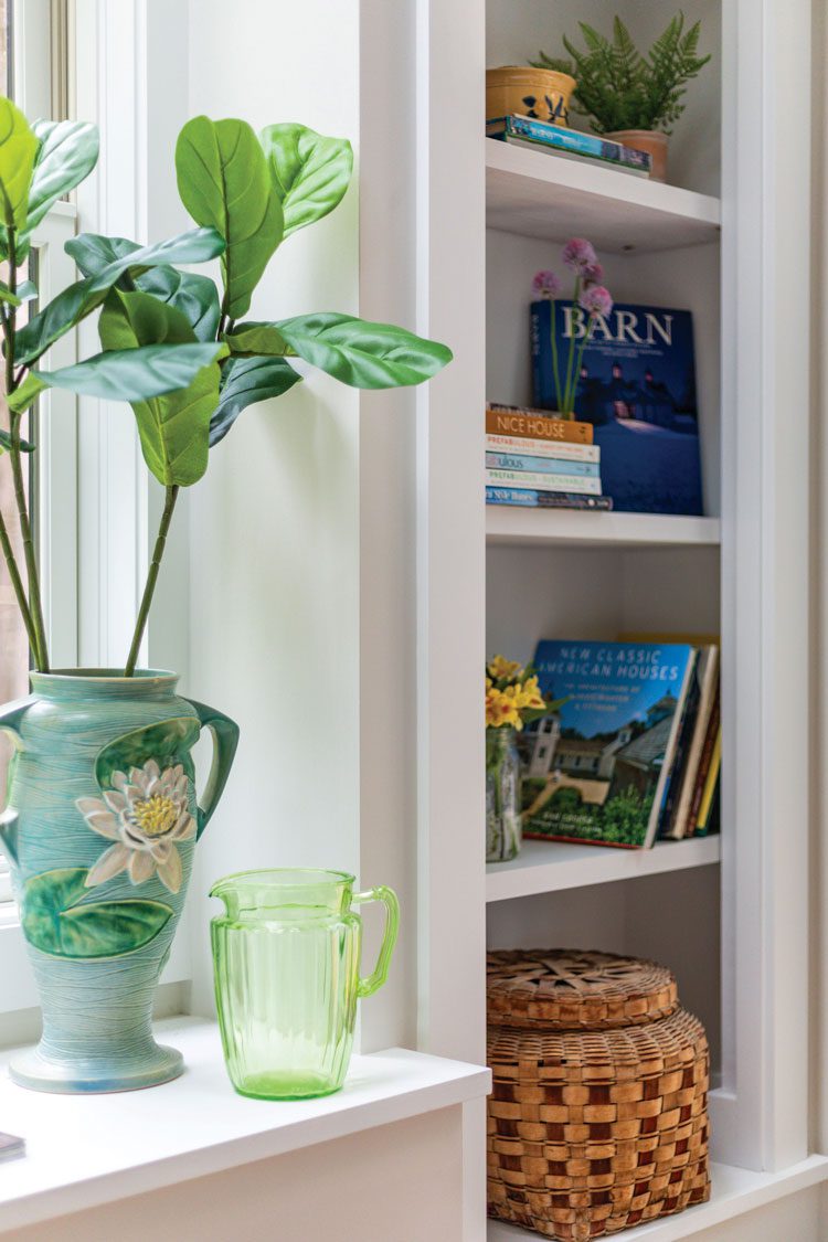 This white, built-in window seat has a blue floral ceramic vase with tall leafy greens and a built-in wall cabinet next to it.