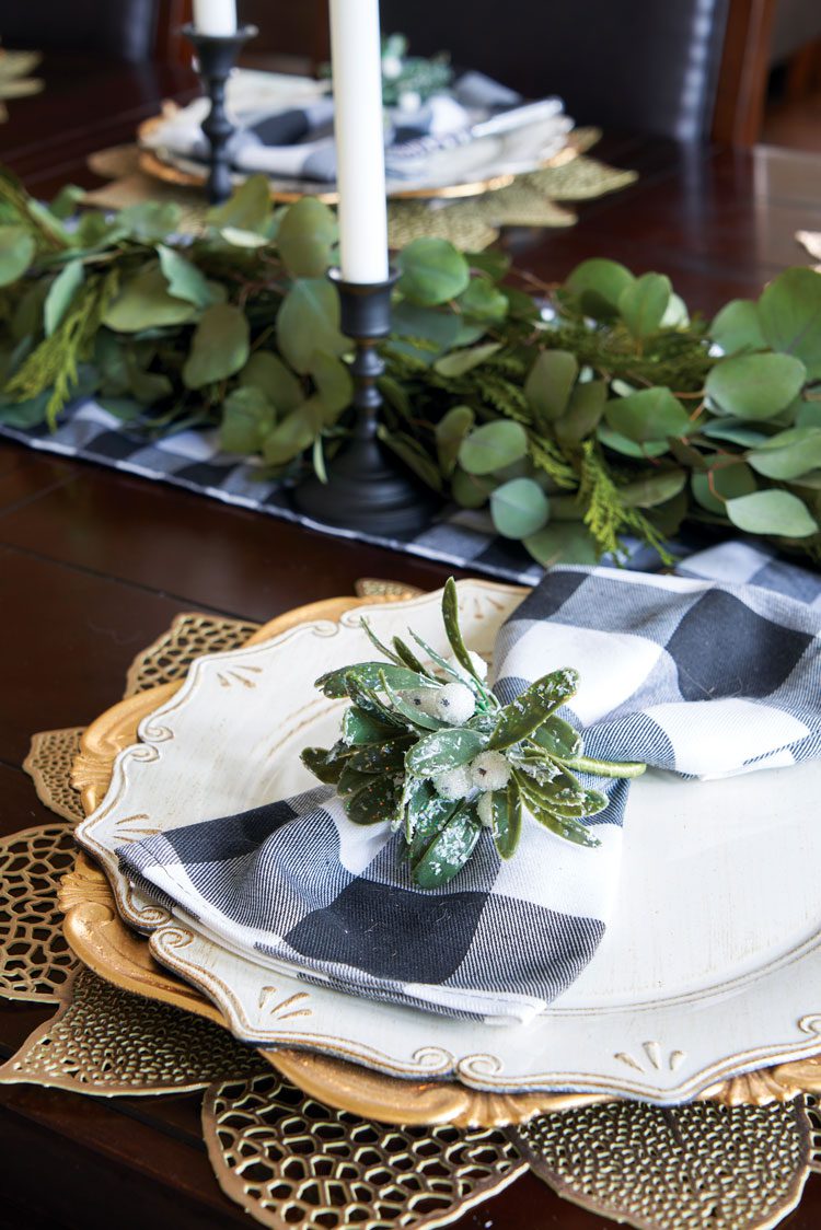 Gold flower place mats beneath a small stack of gold and white dinner plates with a back and white plaid napkin.