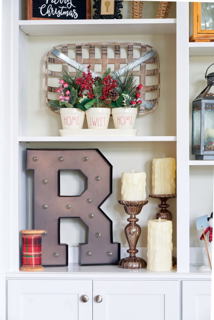 Open shelves in a Christmas bookcase with Rae Dunn pottery filled with faux foliage, candles and a large rusted-looking "R" for Ratliff.