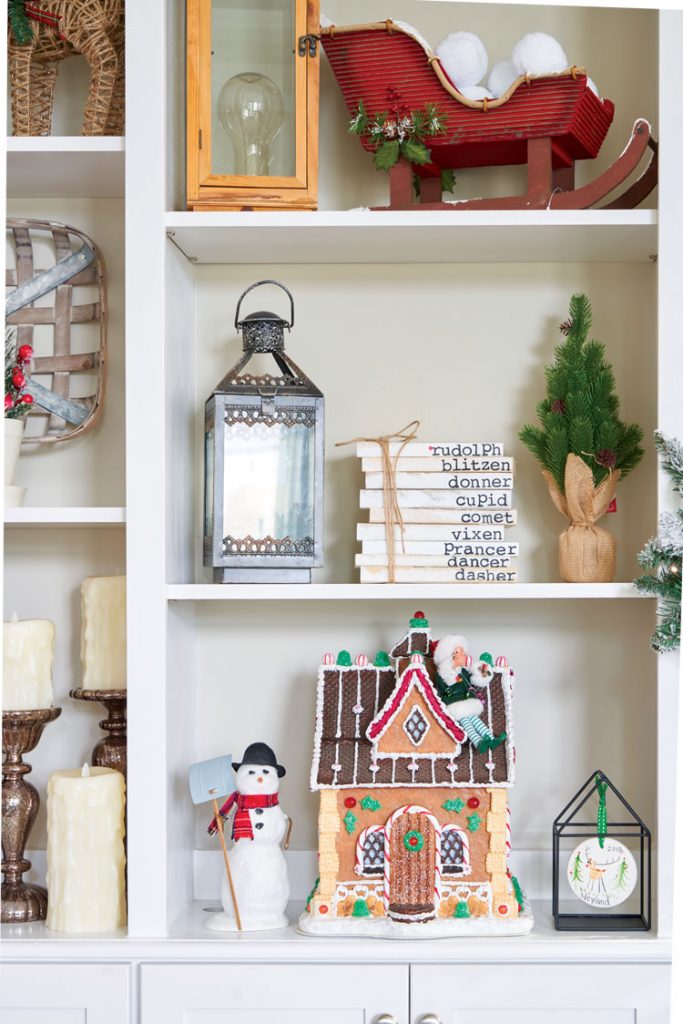 Open shelves in a Christmas bookcase decorated with Christmas decor, including a gingerbread house with an elf-on-the-shelf on the roof.