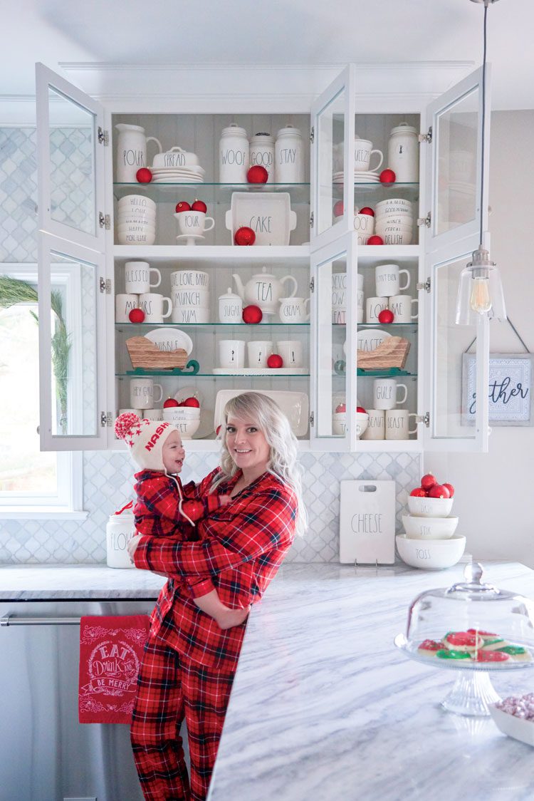 Jenna and her toddler smile in front of her collection of Rae Dunn pottery and red ornaments inside the white kitchen cabinets of her family friendly Christmas house.
