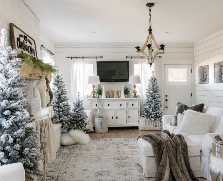 The white Christmas home living room decorated with several snow-crusted pine trees and many pillows and blankets in a neutral color scheme.