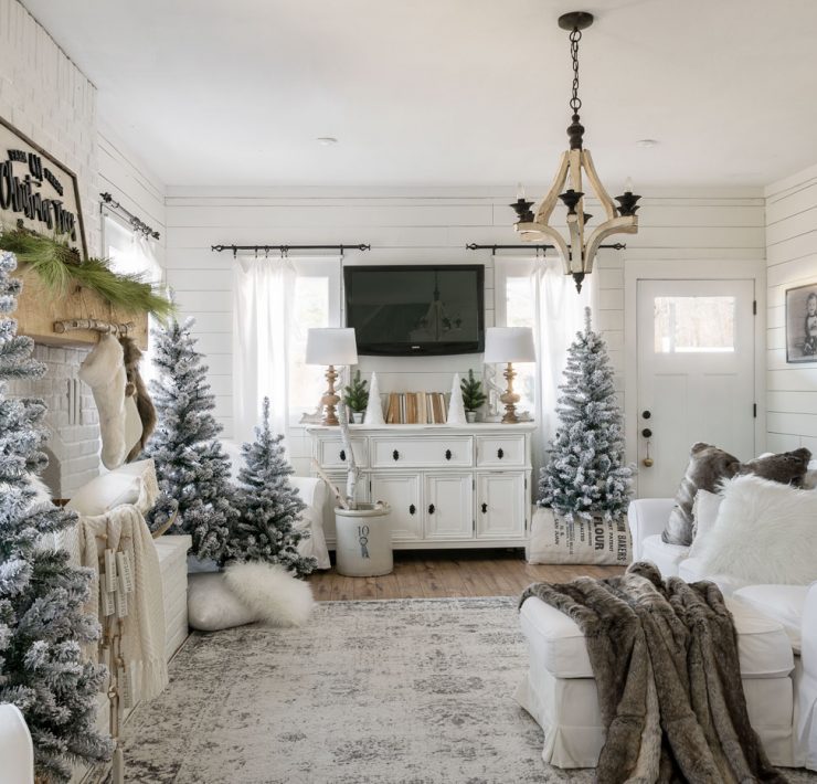 The white Christmas home living room decorated with several snow-crusted pine trees and many pillows and blankets in a neutral color scheme.