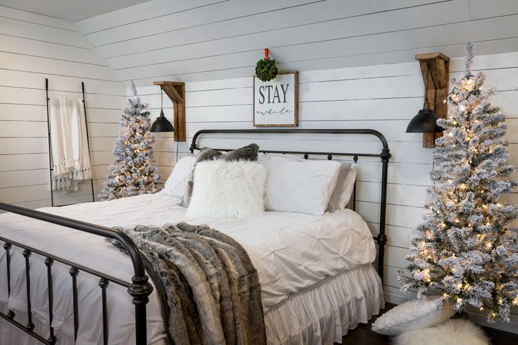 The master bedroom of the white Christmas home with pine trees decorated in faux snow and twinkle lights on either side of the large bed.