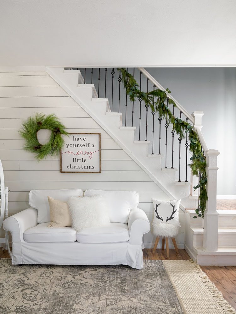 A small, white couch is placed in front of a white shiplap wall beneath the garland-adorned staircase.
