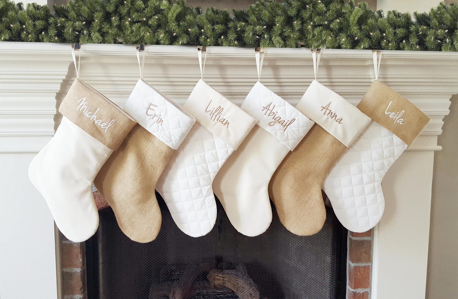 Six personalized stockings in varying neutral shades hanging on a fireplace mantel.