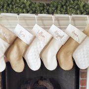 Six personalized stockings in varying neutral shades hanging on a fireplace mantel.