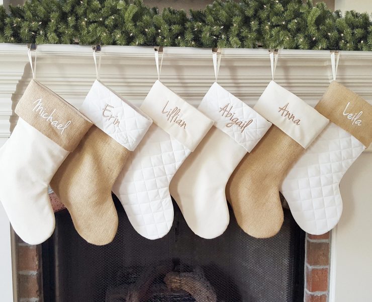 Six personalized stockings in varying neutral shades hanging on a fireplace mantel.