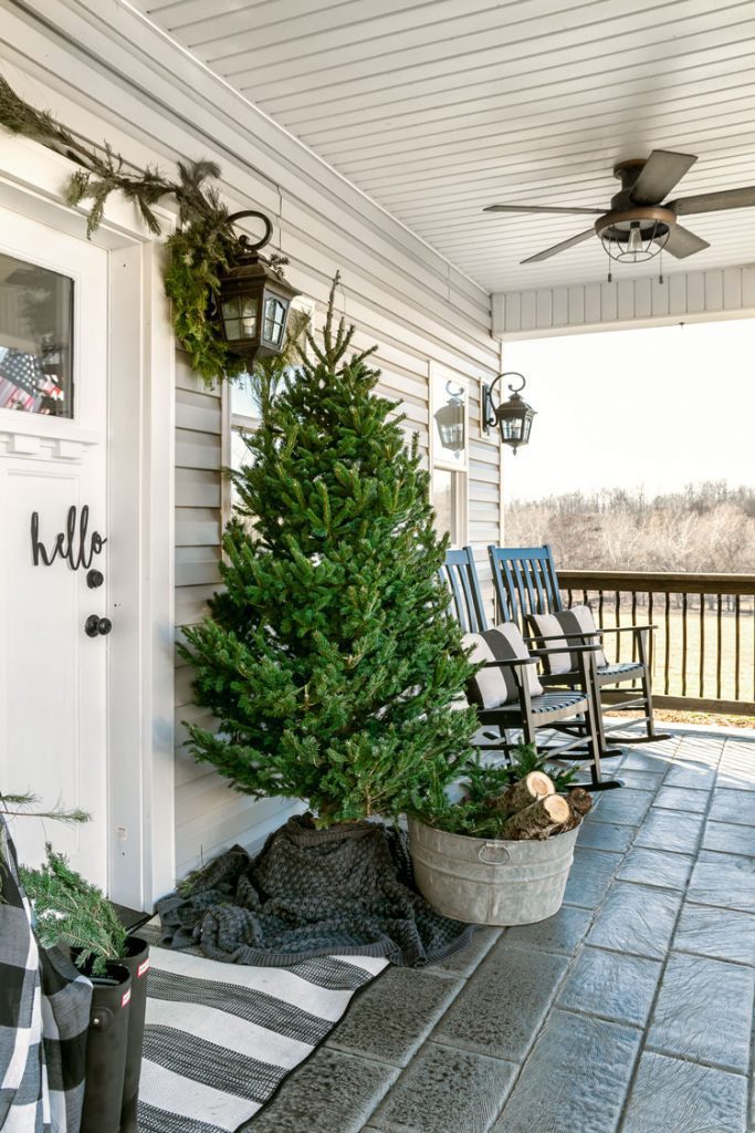 The outside porch of the white Christmas home with stone flooring, a decorative pine tree and a galvanized steel bucket full of chopped wood.
