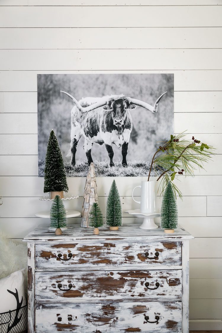 Distressed dresser with sparkly mini pine trees on top. A black and white photo of a cow hangs above it, perfect for a neutral Christmas color scheme.
