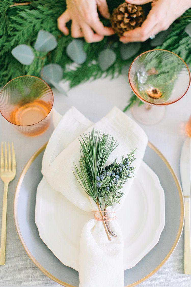 Each place setting has a gold rimmed gray plate beneath a white salad plate with a white linen napkin on top. The napkin is garnished with a spruce tip wrapped in copper wire.