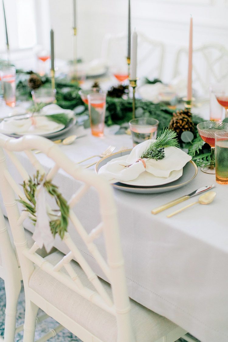 Christmas dinner table set with white chairs, tablecloth and dishes, accented by evergreen, candlesticks and mixed metals.