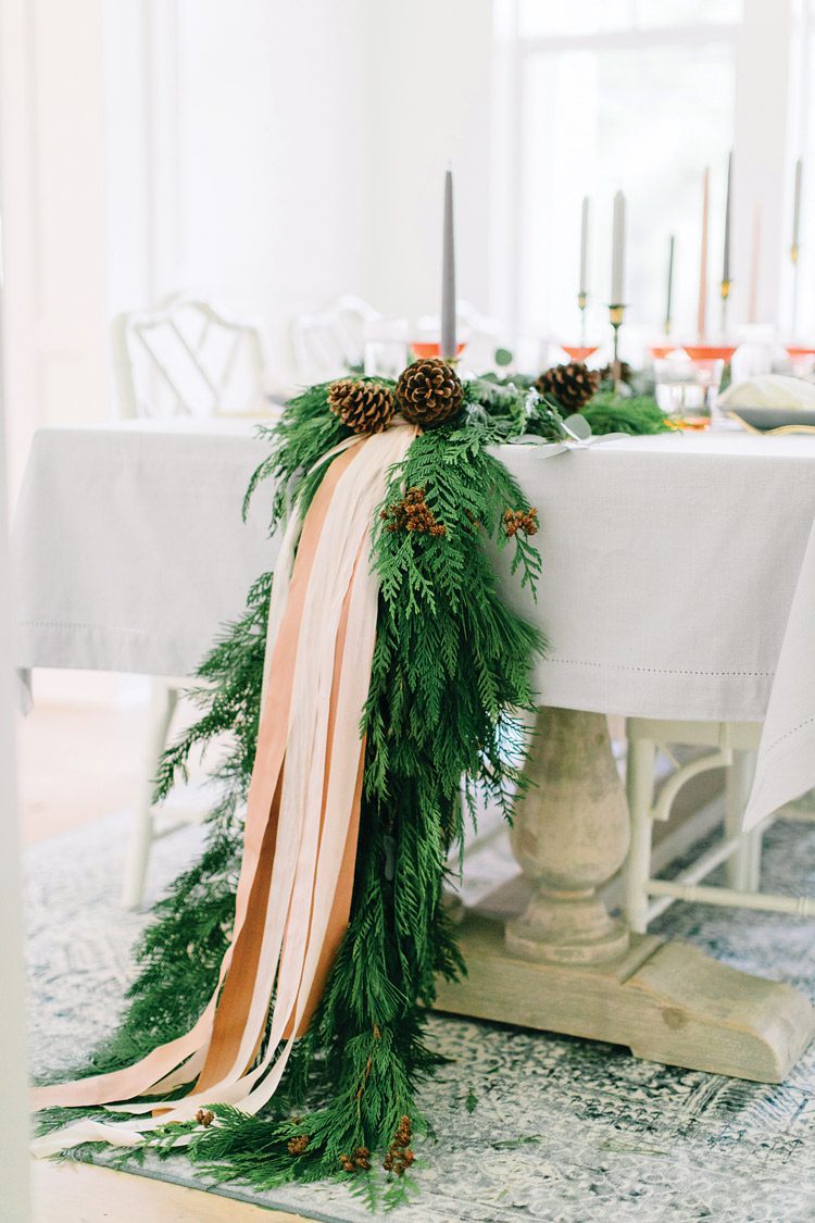 The evergreen garland droops down the side of the Christmas dinner table and onto the floor, along with muted pink ribbons.