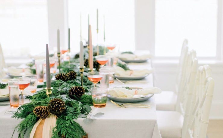 The Christmas dinner table has lots of natural light from the windows and a large evergreen garland running down the middle of it.