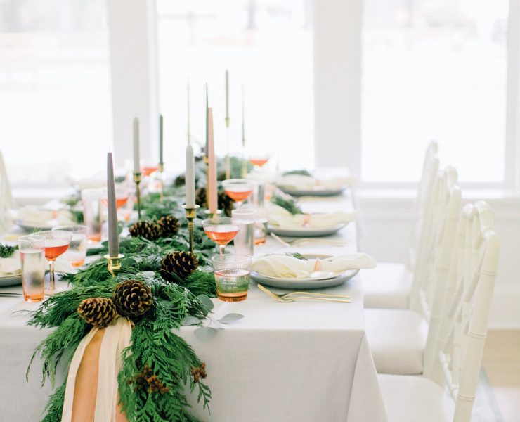 The Christmas dinner table has lots of natural light from the windows and a large evergreen garland running down the middle of it.