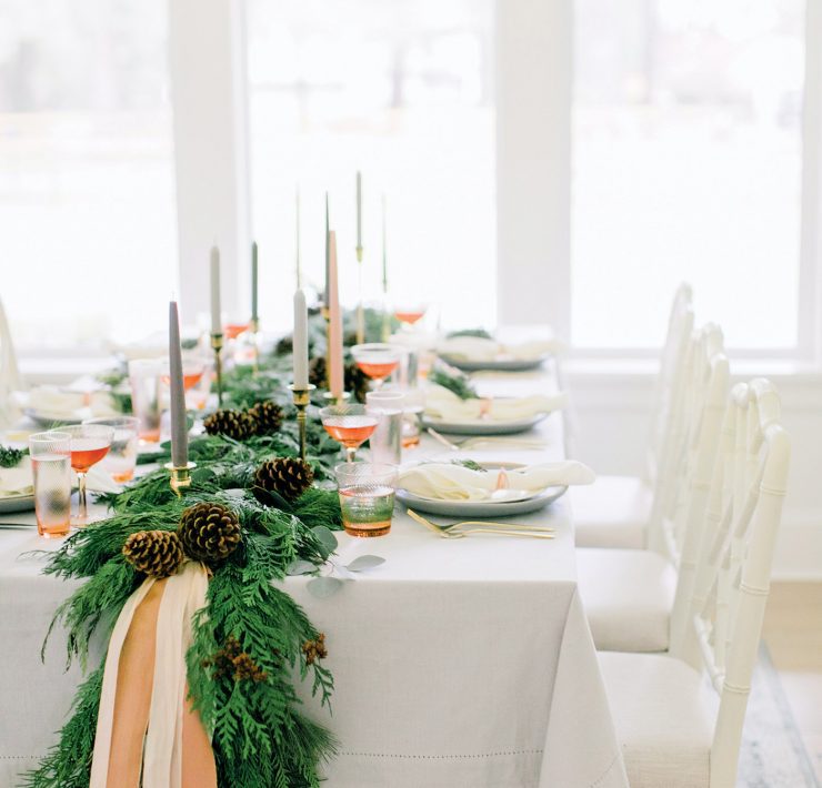 The Christmas dinner table has lots of natural light from the windows and a large evergreen garland running down the middle of it.