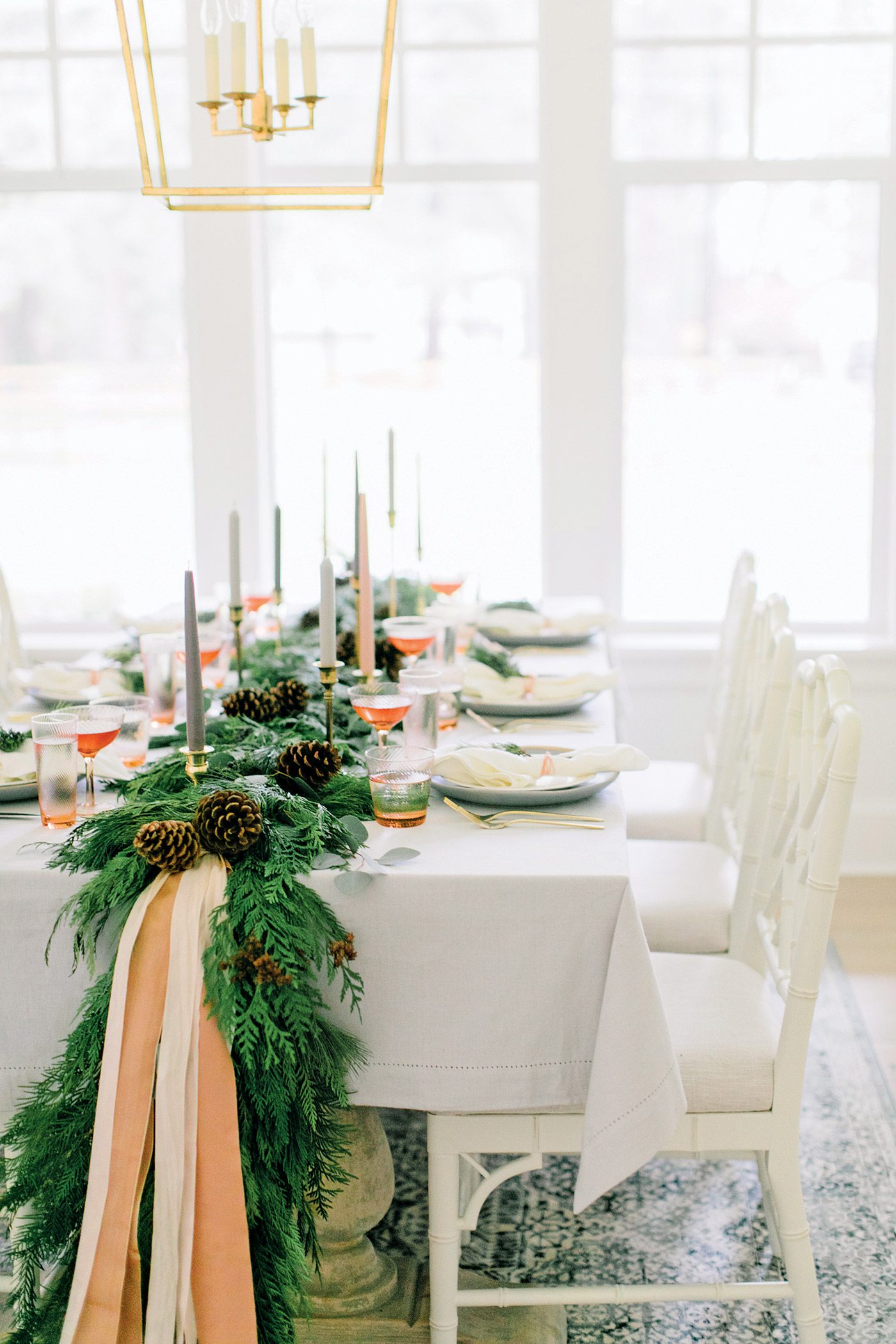 The Christmas dinner table has lots of natural light from the windows and a large evergreen garland running down the middle of it.
