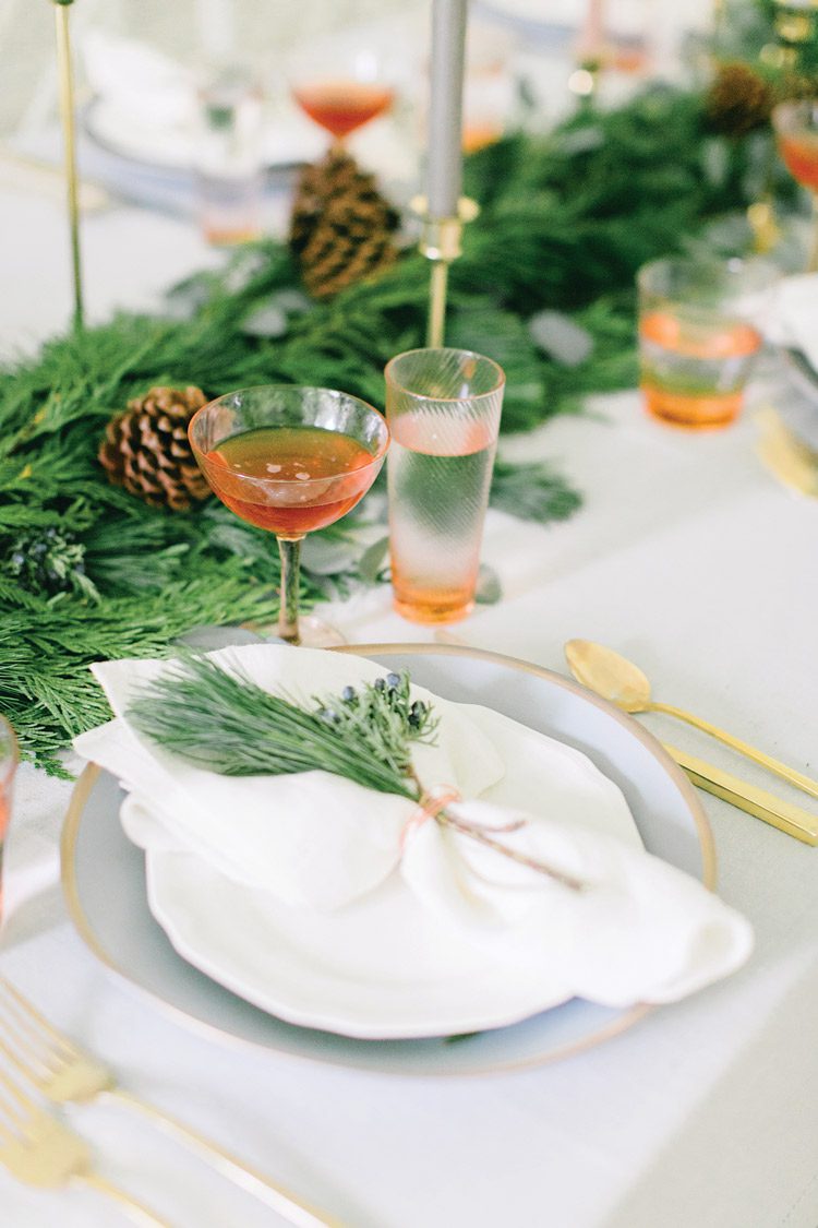 Stemmed and regular glasses on the right side of a gray and white place setting with an evergreen garland running down the middle of the Christmas dinner table.