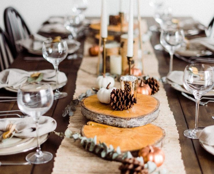 This fall tablescape has a burlap runner decorated with large wood slices, pinecones, seasonal greens, small pumpkins and candlesticks.