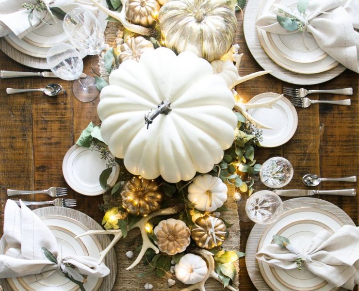 This design is a glamorous take on fall tables capes. It has a burlap runner over a bare wood table, decorated with small and larges pumpkins spray-painted white, gold and silver. It also has tiny faux antlers dispersed throughout.