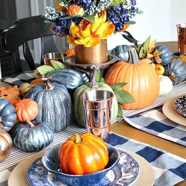 The brightest of our fall tablescapes round up, this design has orange and blue-painted pumpkins piled along the center with patterned blue dishes, place mats and silver cups.