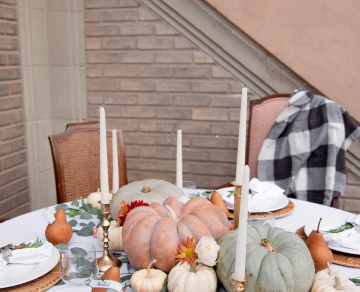 This is the lightest color palette among our fall tablescapes round up. It is a circular table with a white tablecloth and plates, with a bronze-painted pear bearing each person's name. In the center of the table, tall candlesticks are placed around a decorative display of off-white, pastel green and orange pumpkins.