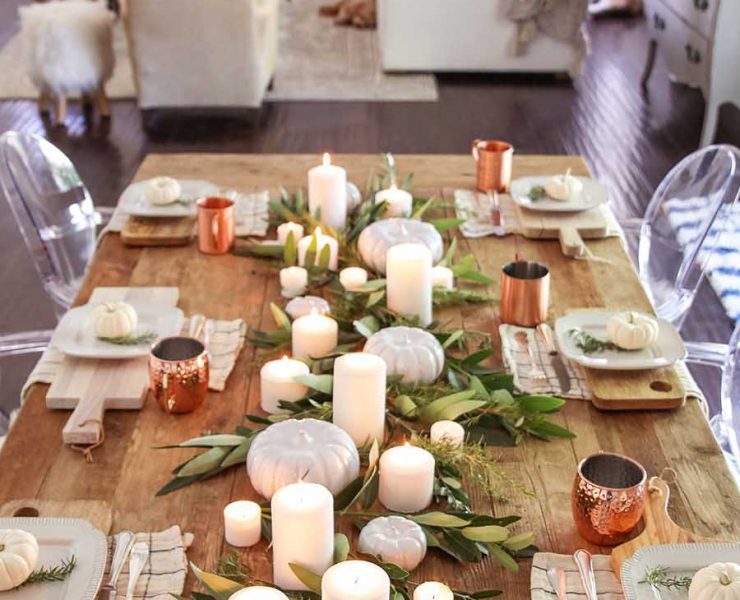 A farmhouse minimalist design among fall tablescapes, this design features wood cutting boards as place mats, copper mugs, and a center display of white candles, white pumpkins and leafy greens.