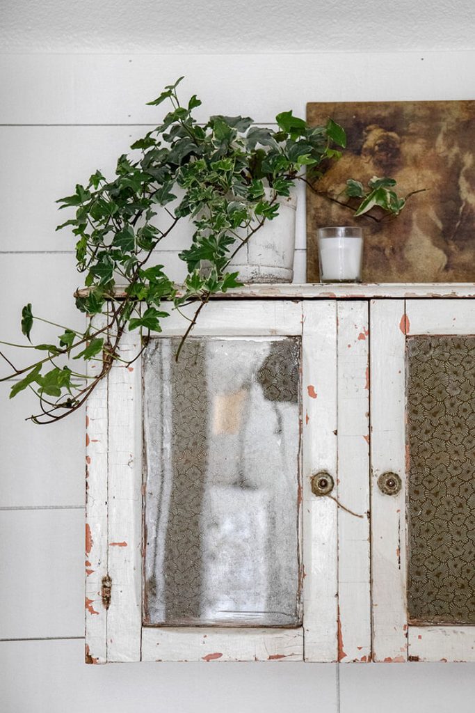 A chippy closed cabinet hanging from the wall with ivy planter on top